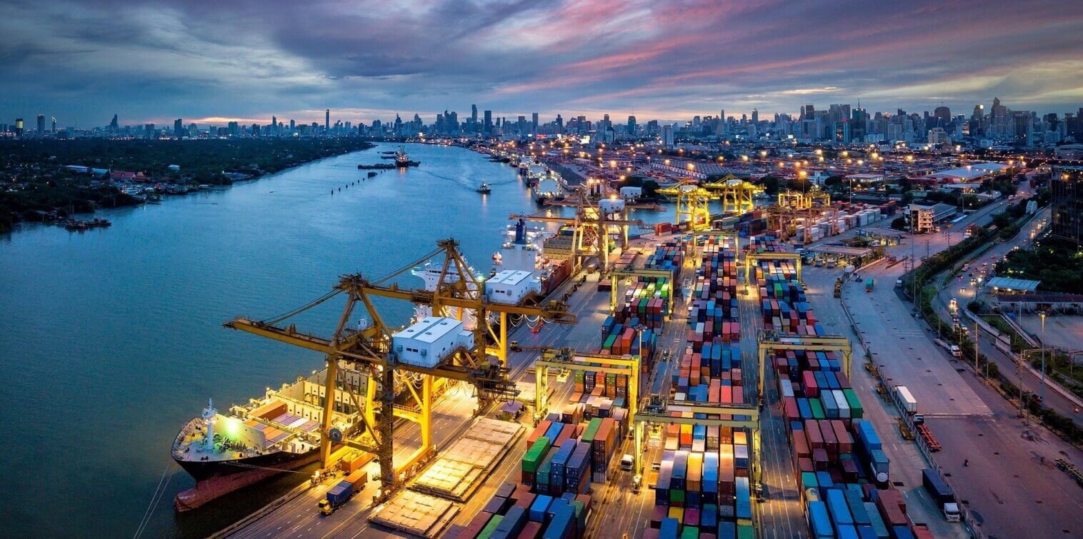 Aerial view of the Port of Hamburg at dusk, with container terminals, cargo ships, cranes and the Hamburg skyline illuminated in the background.