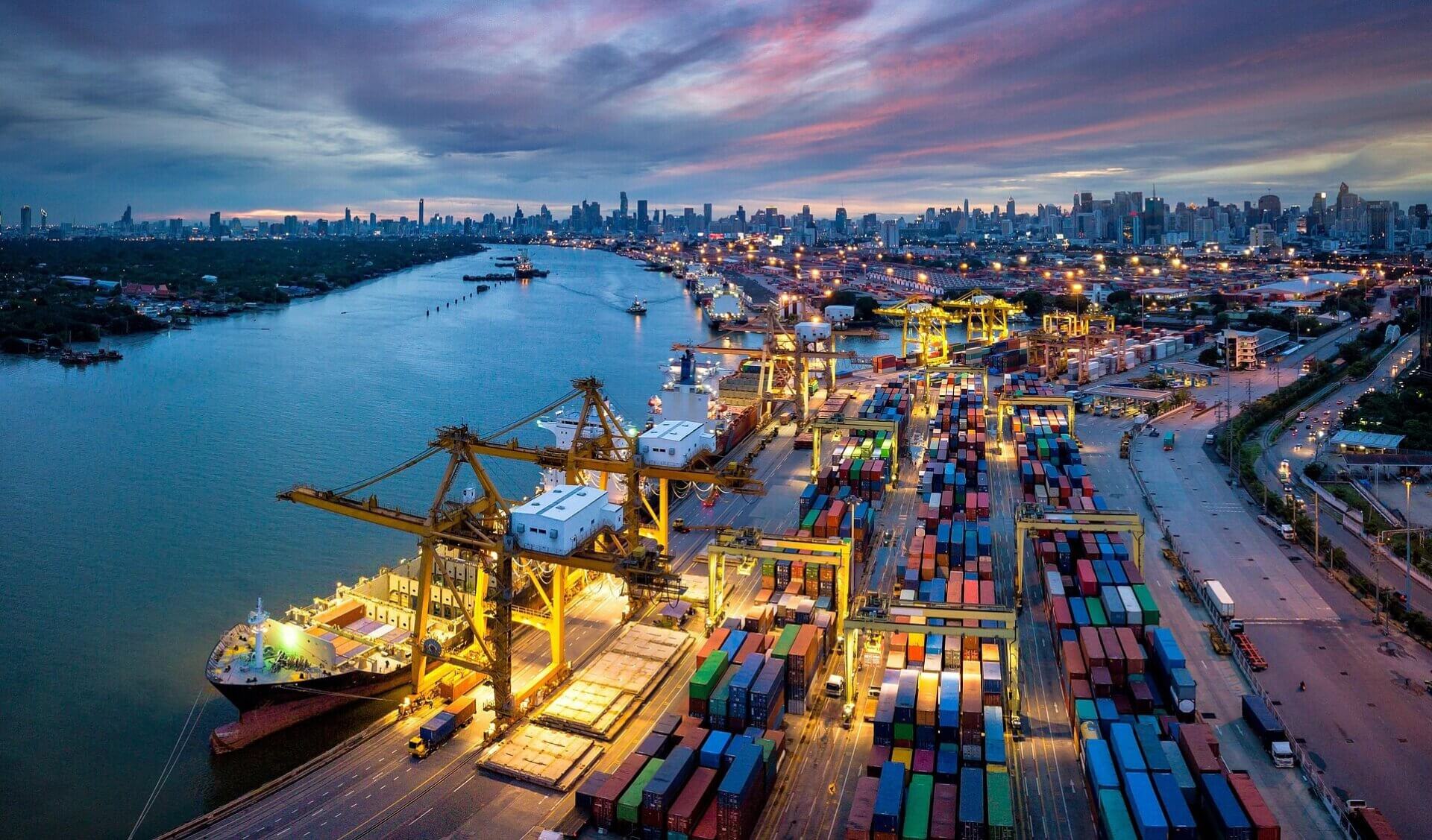 Aerial view of the Port of Hamburg at dusk, with container terminals, cargo ships, cranes and the Hamburg skyline illuminated in the background.